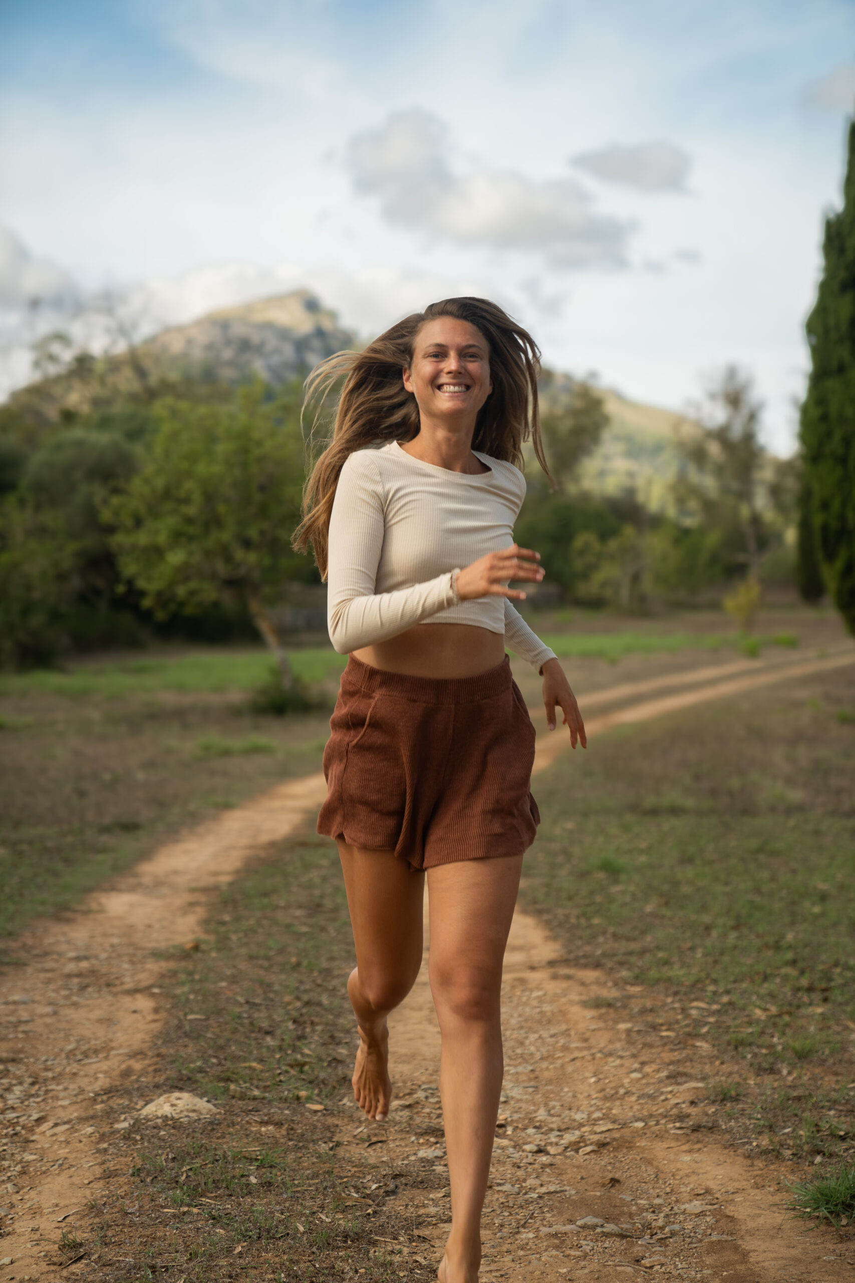 Ein Foto von Jana Buth, wie sie auf einem Weg in der Natur läuft. Sie joggt auf die Kamera zu und lächelt dabei. Sie trägt eine rote kurze Jeans und ein weißes langärmliges Shirt. Im Hintergrund sieht man Bäume und Berge.