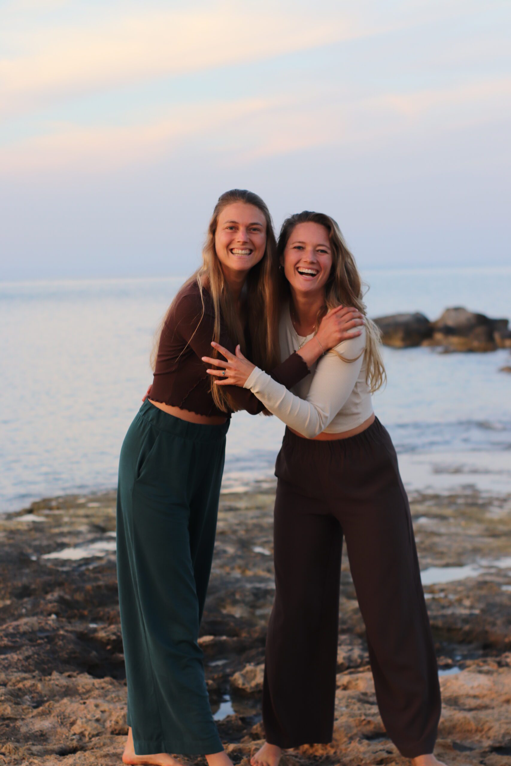 Auf dem Foto stehen Jana und Kristin vor dem Meer und umarmen sich. Sie schauen beide zur Kamera und lächeln.